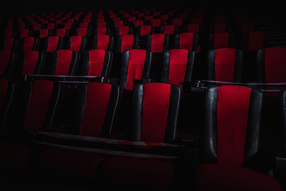 Poltrone vuote da cinema, con cuscini centrali rossi e struttura nera, disposte in file in una sala scarsamente illuminata.