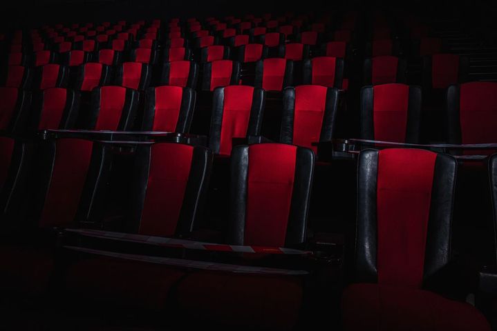 Poltrone vuote da cinema, con cuscini centrali rossi e struttura nera, disposte in file in una sala scarsamente illuminata.