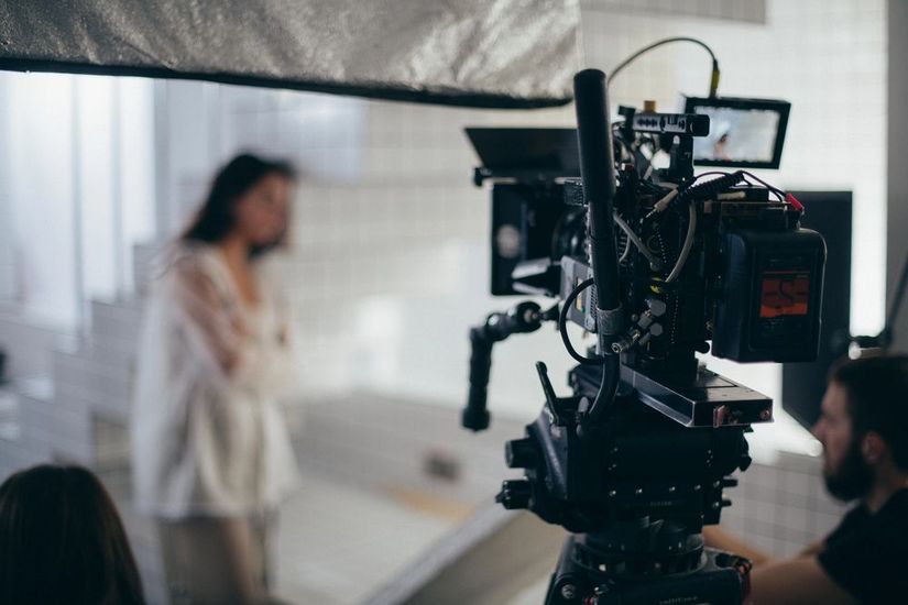 Una cinepresa professionale su treppiede è puntata verso una persona in piedi in uno studio fotografico illuminato a giorno e con l'immagine sfocata.
