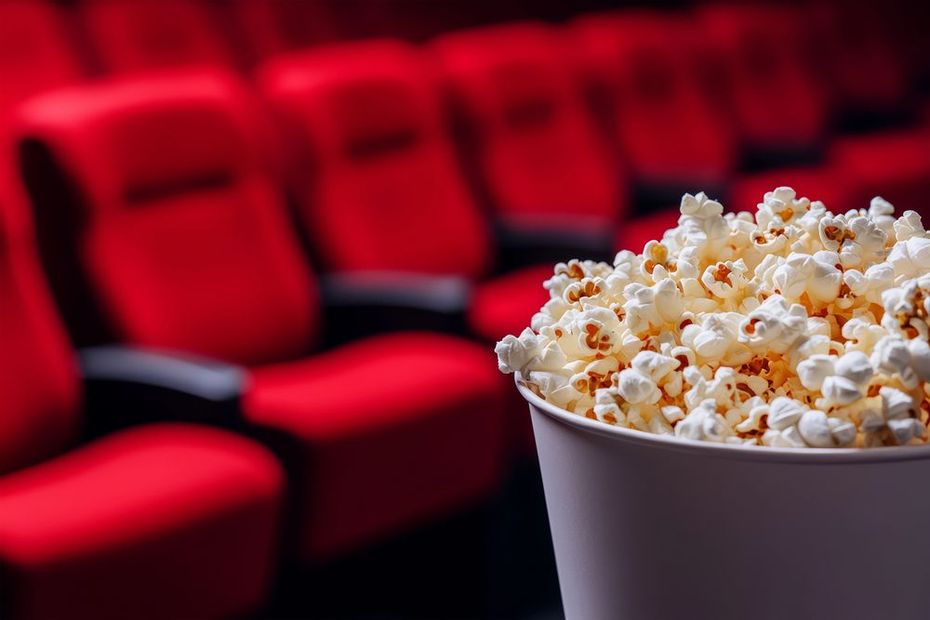 In primo piano un grande secchio di popcorn, con sullo sfondo file di poltrone da cinema vuote e sfocate di colore rosso.