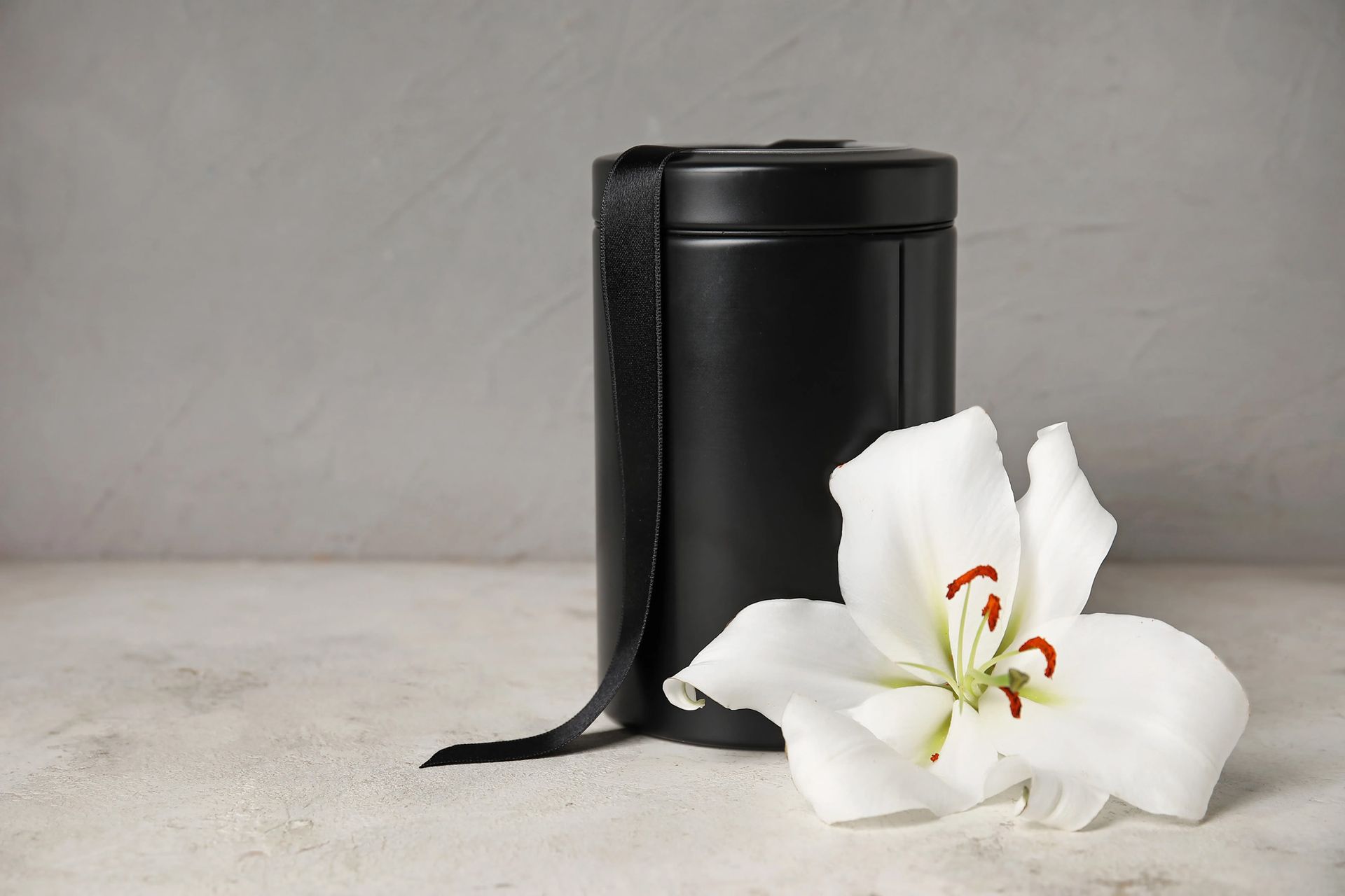 Black urn with a black ribbon and white lily on a light surface against a gray background.
