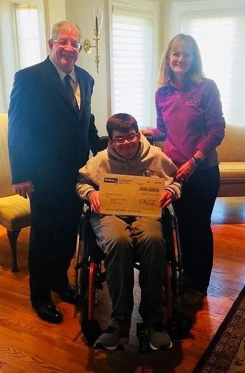 Man in wheelchair holding check, flanked by two people in a room, receiving donation.