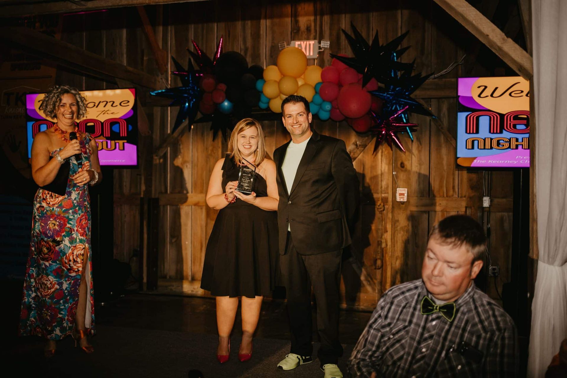 People at an event pose for a photo. A woman in a floral dress speaks into a microphone. A couple smiles, with the woman holding an award. The man is in a suit.