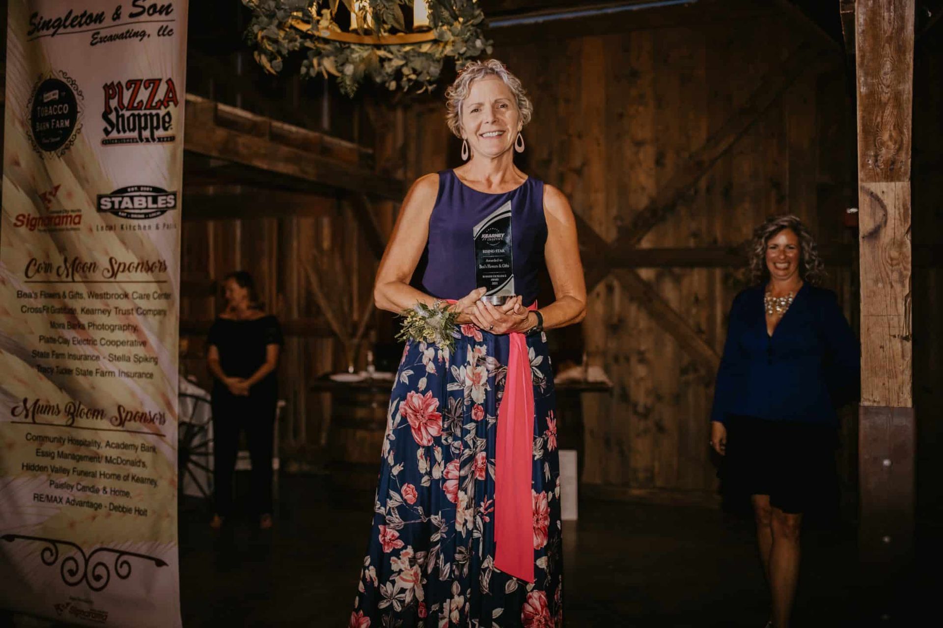 Woman in floral dress holding an award, smiling, in a barn setting with two other women.