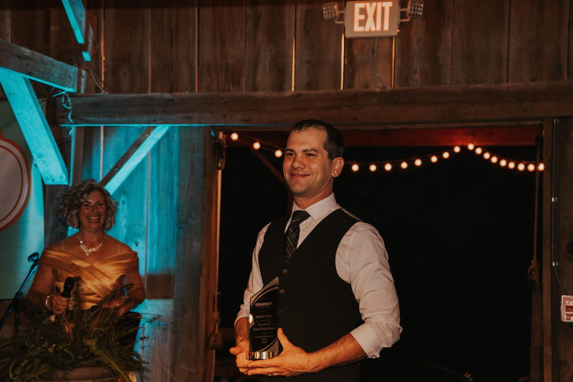 Man in vest holding award, smiling. Woman in gold dress behind him. Inside a barn-like structure with lights.