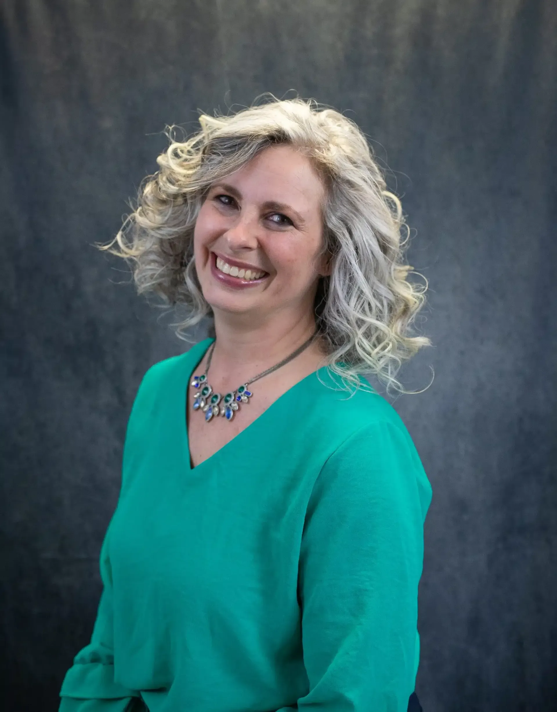 Woman with curly gray hair smiles, wearing a teal sweater and a silver necklace. Gray background.