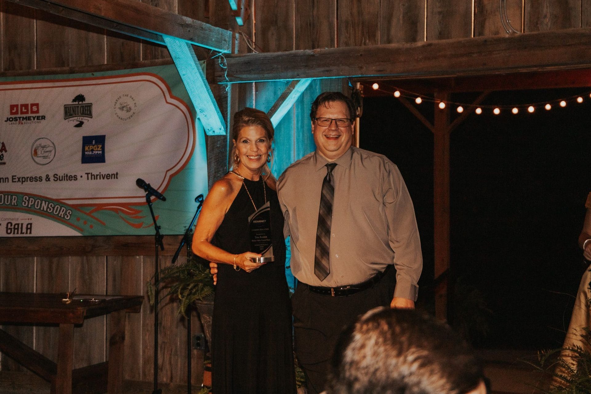 A woman in a black dress and a man in a shirt and tie stand together, smiling, at an event in a barn.