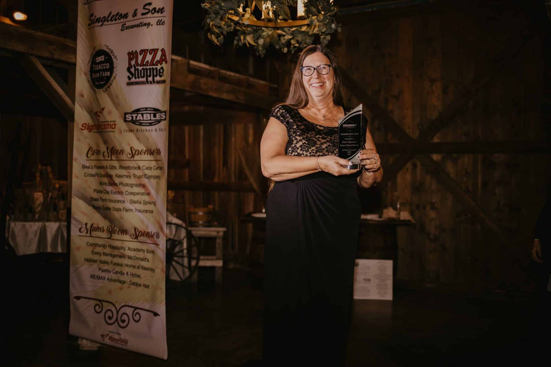 Woman in a black formal gown holding an award in a rustic barn setting, smiling at the camera.