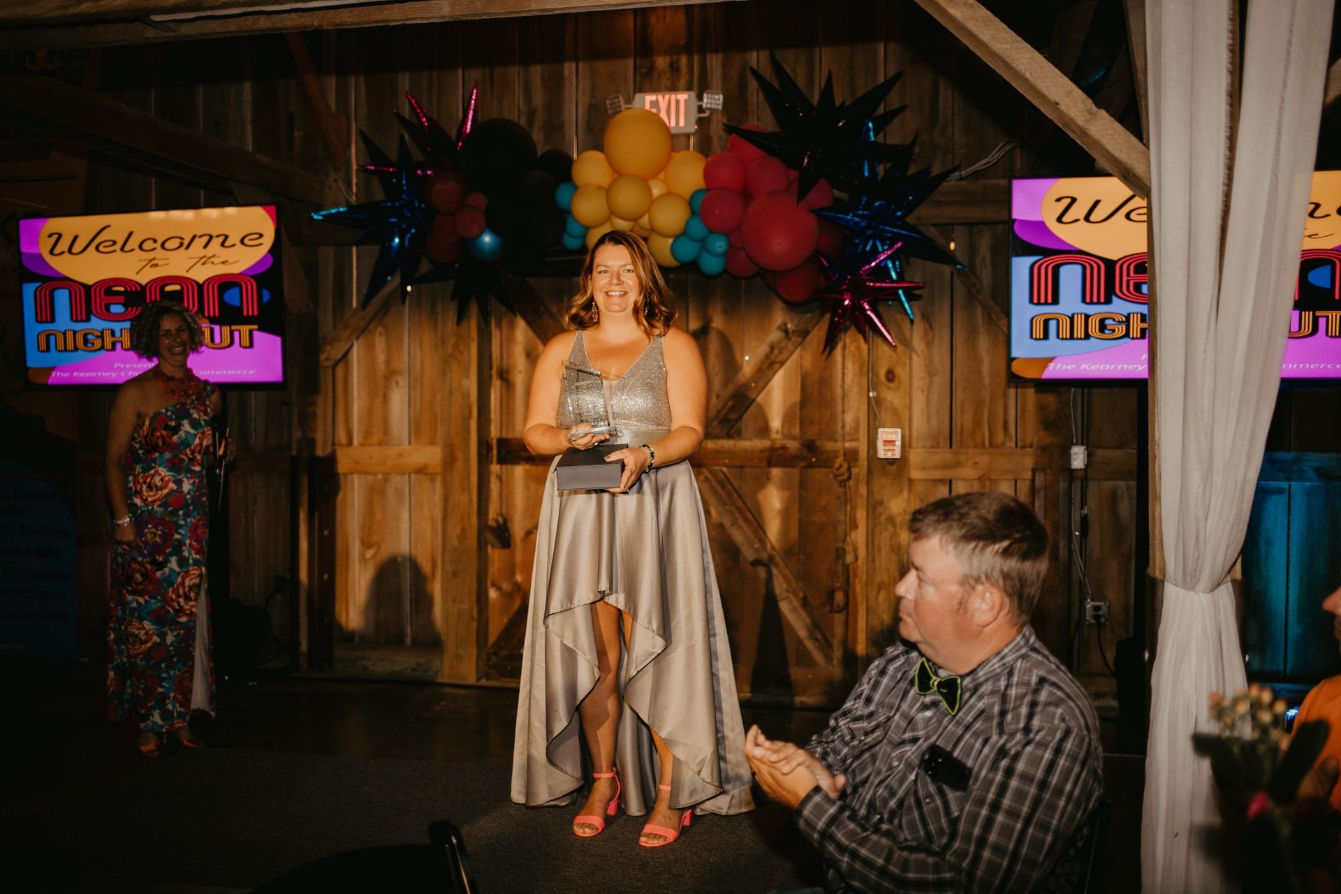 Woman in silver gown accepting an award onstage at an event in a barn; balloons and screens in the background, man applauding.