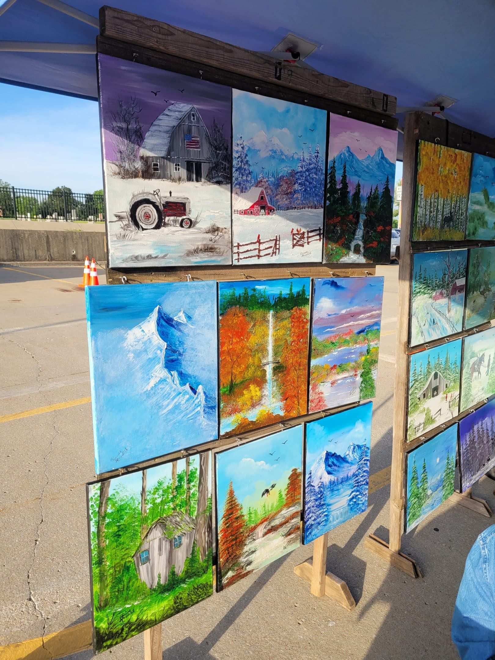 Paintings displayed on a booth. Landscapes feature mountains, forests, and cabins, using a variety of colors.