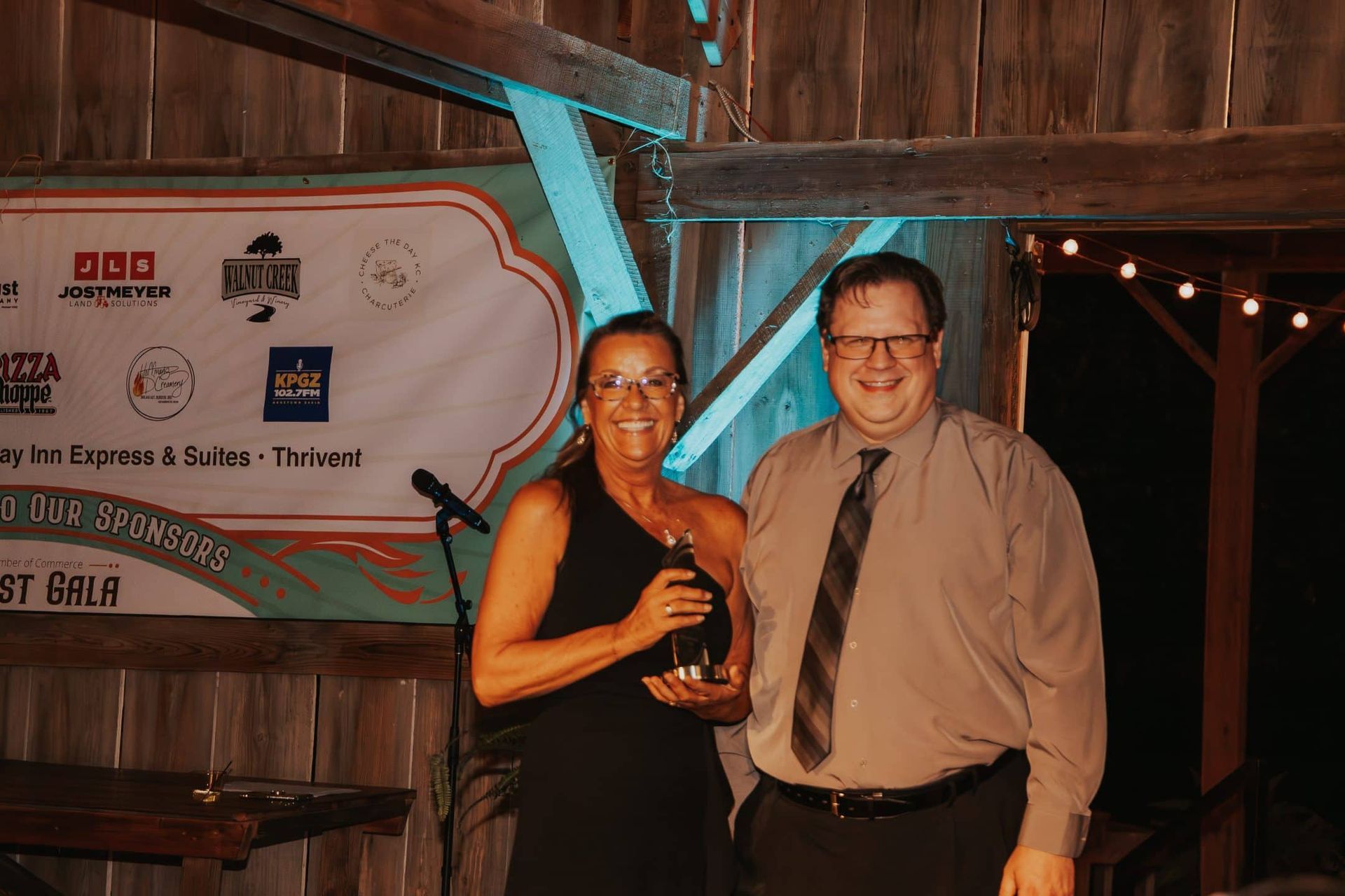 Woman in black dress and man in tan shirt stand together at a gala, the woman holding an award. They are in a barn-like setting.