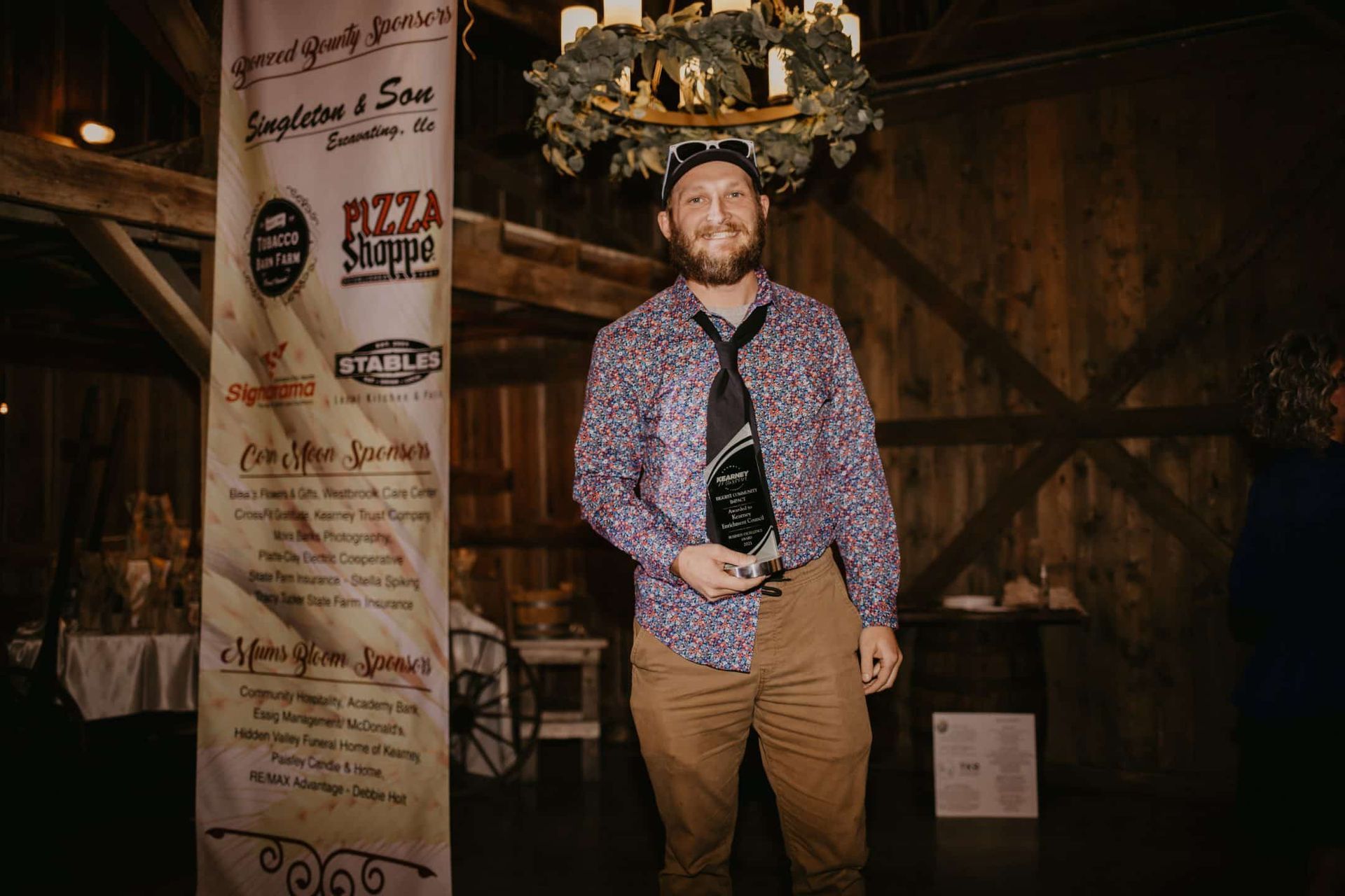 A man in a patterned shirt and a tie holds an award in a barn. He smiles, and a banner stands next to him.