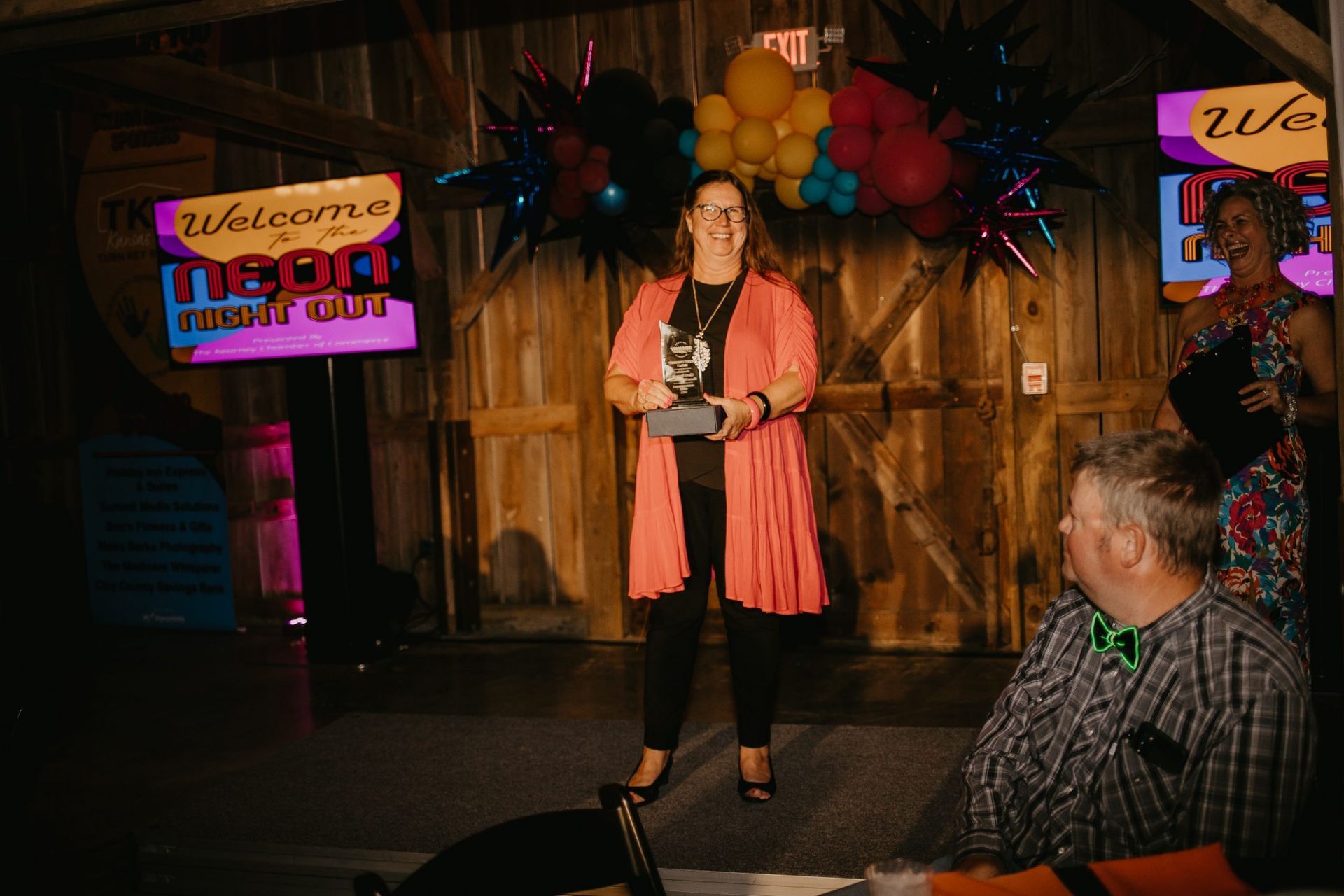 A woman with a trophy stands on a stage in a barn, smiling. Balloons and