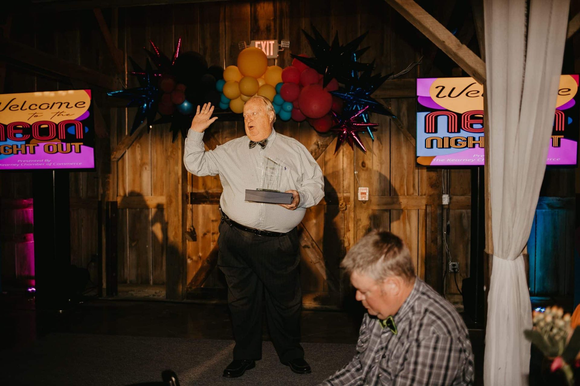 Man waving at an event, holding a tray. Stage is decorated with balloons, signs that read “Welcome Night Out”, and a man seated to the lower right.