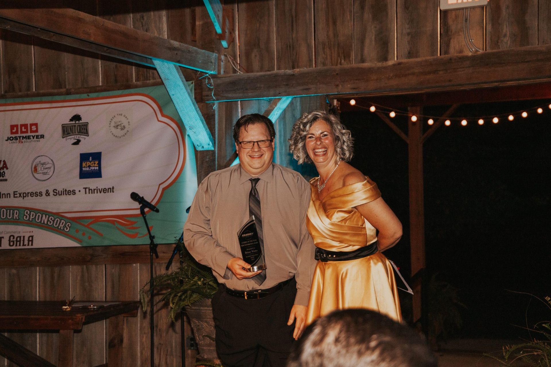 A man in a shirt and tie holds an award next to a woman in a gold dress. They are on a stage with a banner behind them.