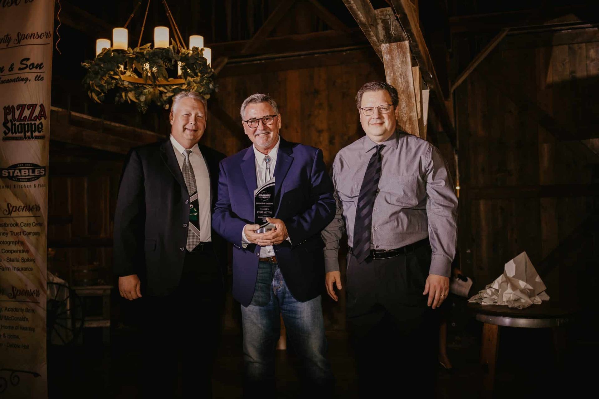 Three men in suits and ties stand indoors, one holding an award. The setting is dimly lit with rustic decor.