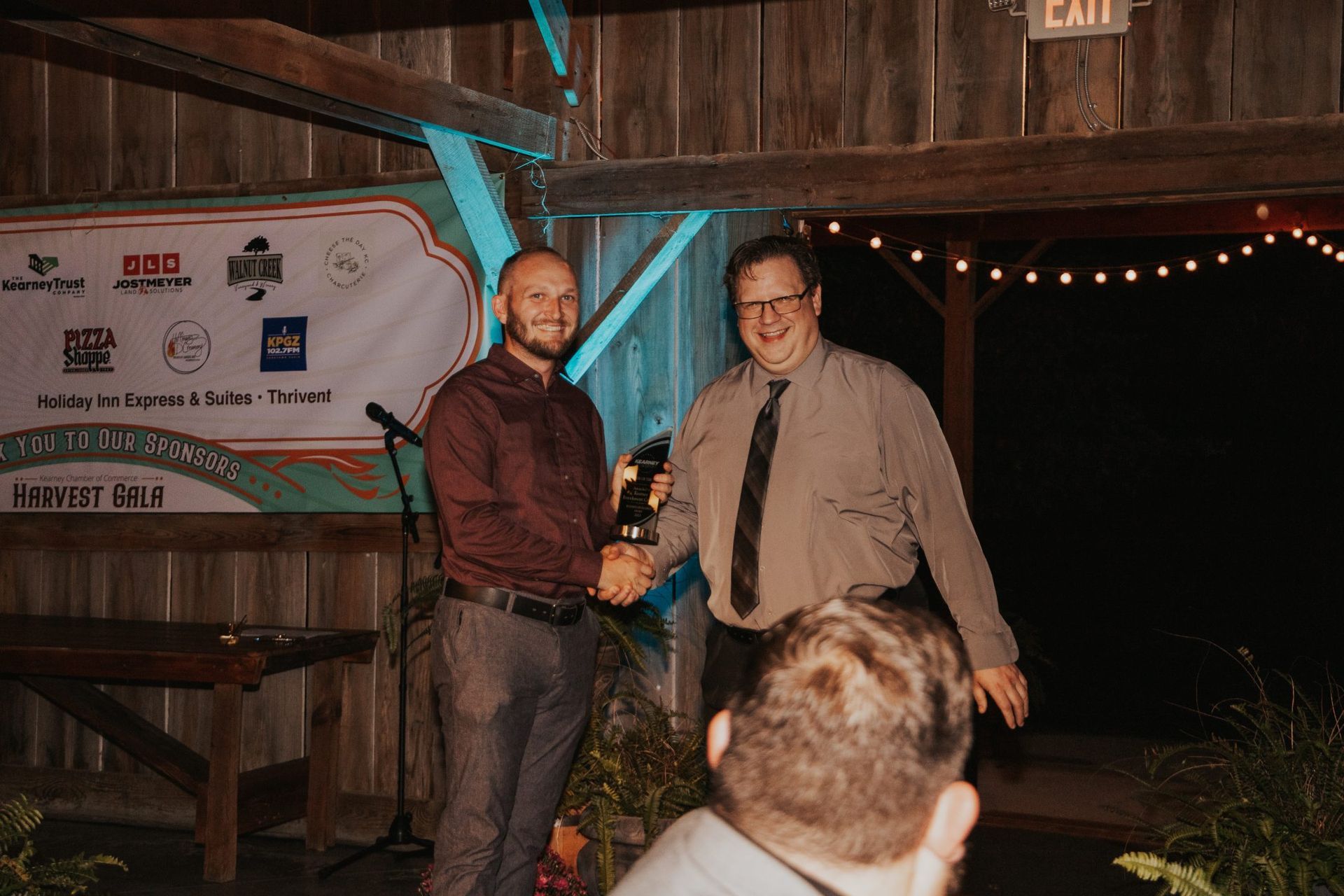 Two men shaking hands at an event. One man is wearing a maroon shirt and the other a grey shirt and tie. They stand in front of a banner in a wooden building.