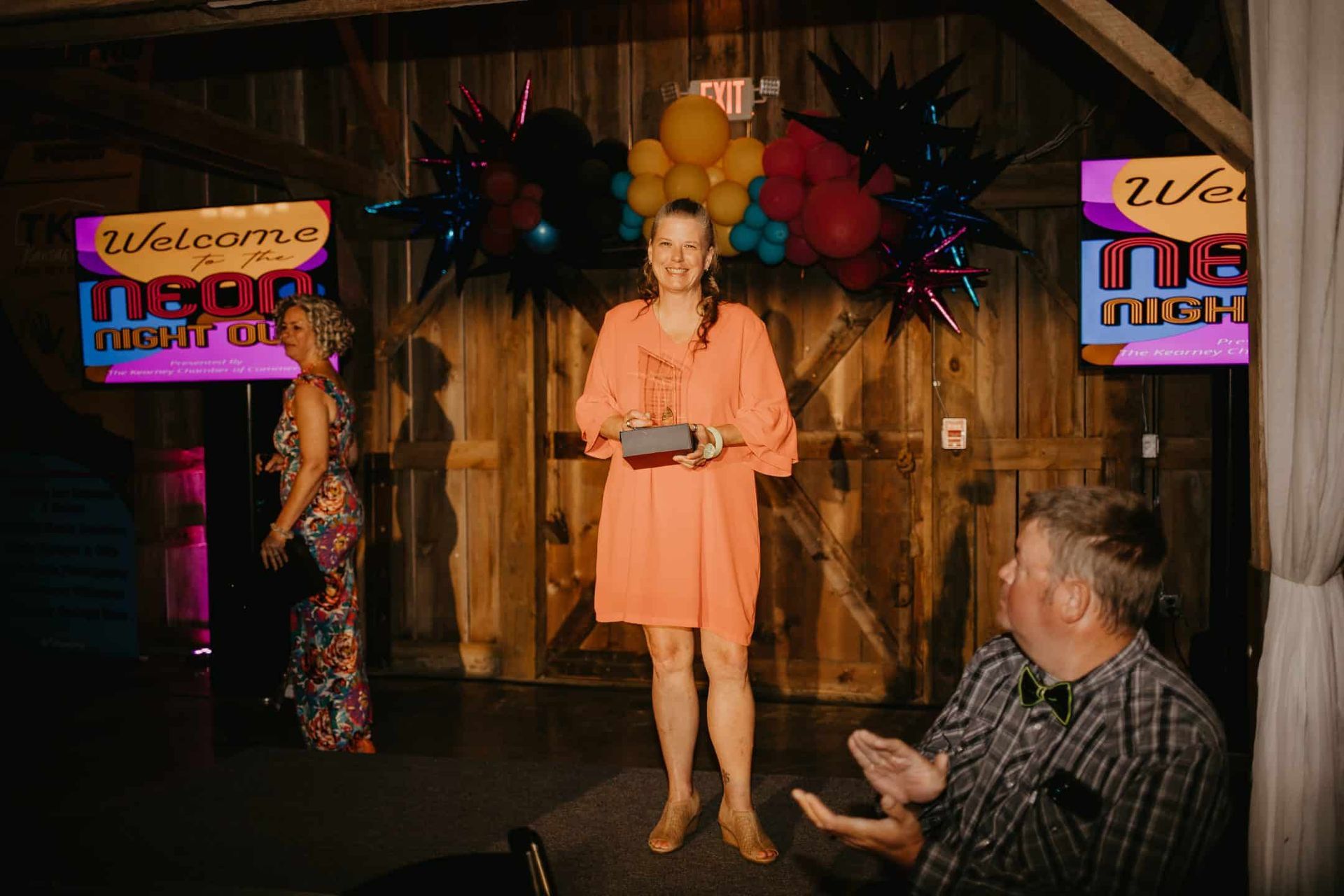 Woman in orange dress holding an award, standing in front of a backdrop. A woman in a patterned dress walks past, and a man applauds.
