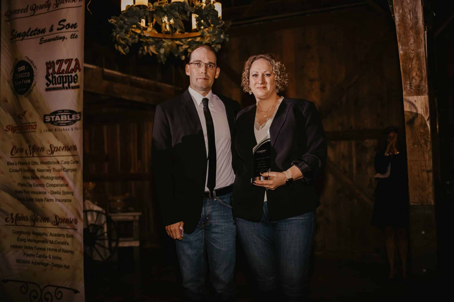 A man and woman stand close together inside a rustic building. The man wears a suit and tie, the woman a dark jacket.