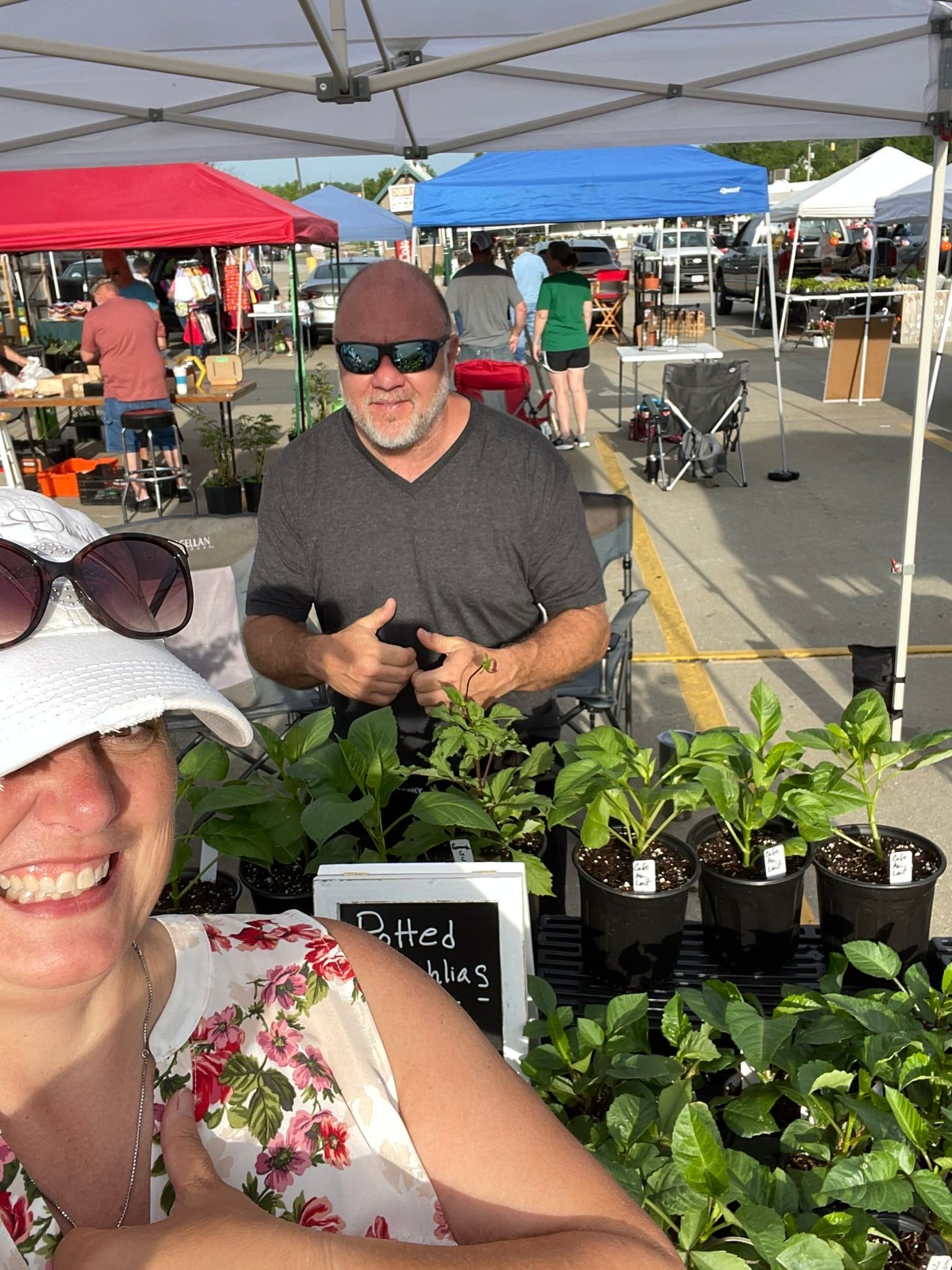 A woman and a man smile at a plant stand at an outdoor market. The man gives a thumbs-up.