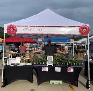 A man stands behind a flower stall at an outdoor market under a white canopy tent. He is surrounded by flowers and plants.