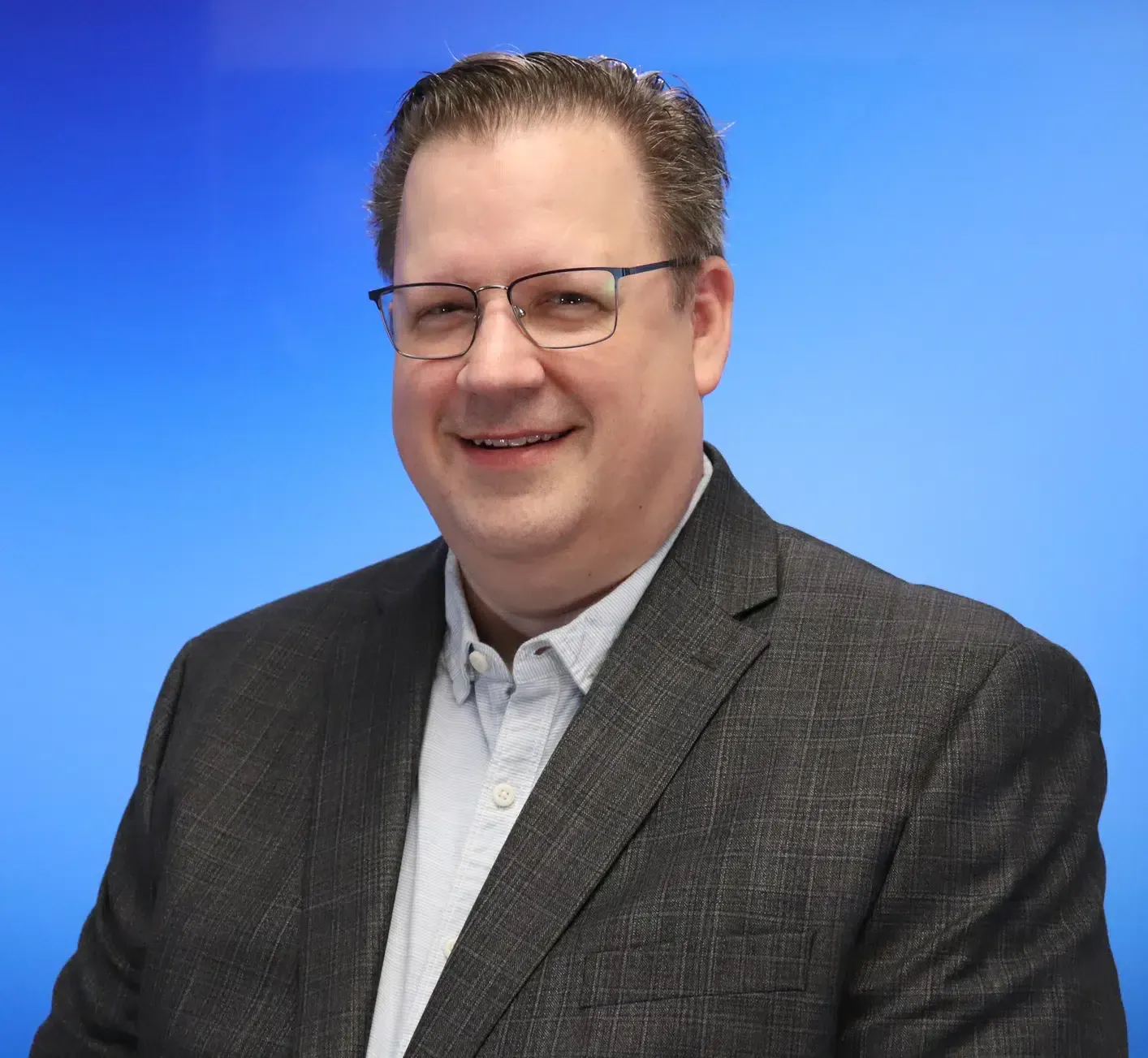 A smiling man with glasses in a dark blazer and button-down shirt against a blue gradient background.