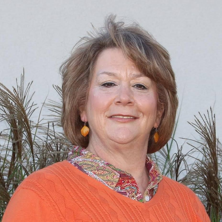 Woman with shoulder-length, light brown hair, wearing an orange sweater and patterned shirt smiling outdoors with neutral background and tall grass.