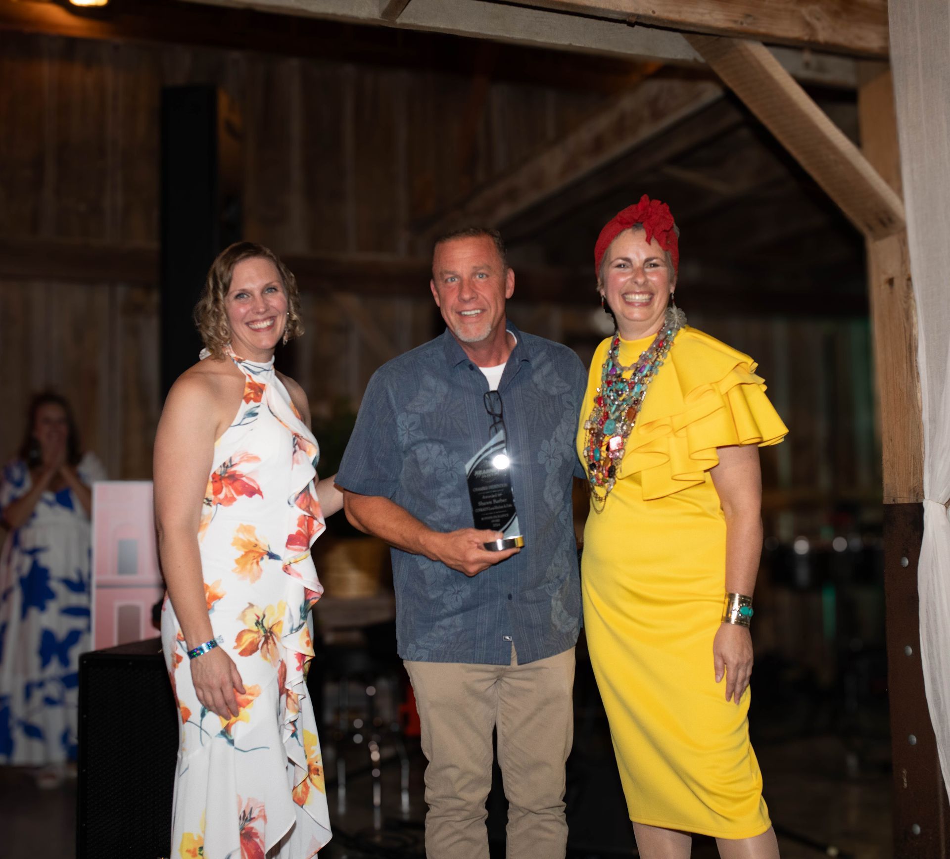 Three people posing for a photo at an event; a man holding an award between two women, all smiling. The setting appears to be a barn-like structure.