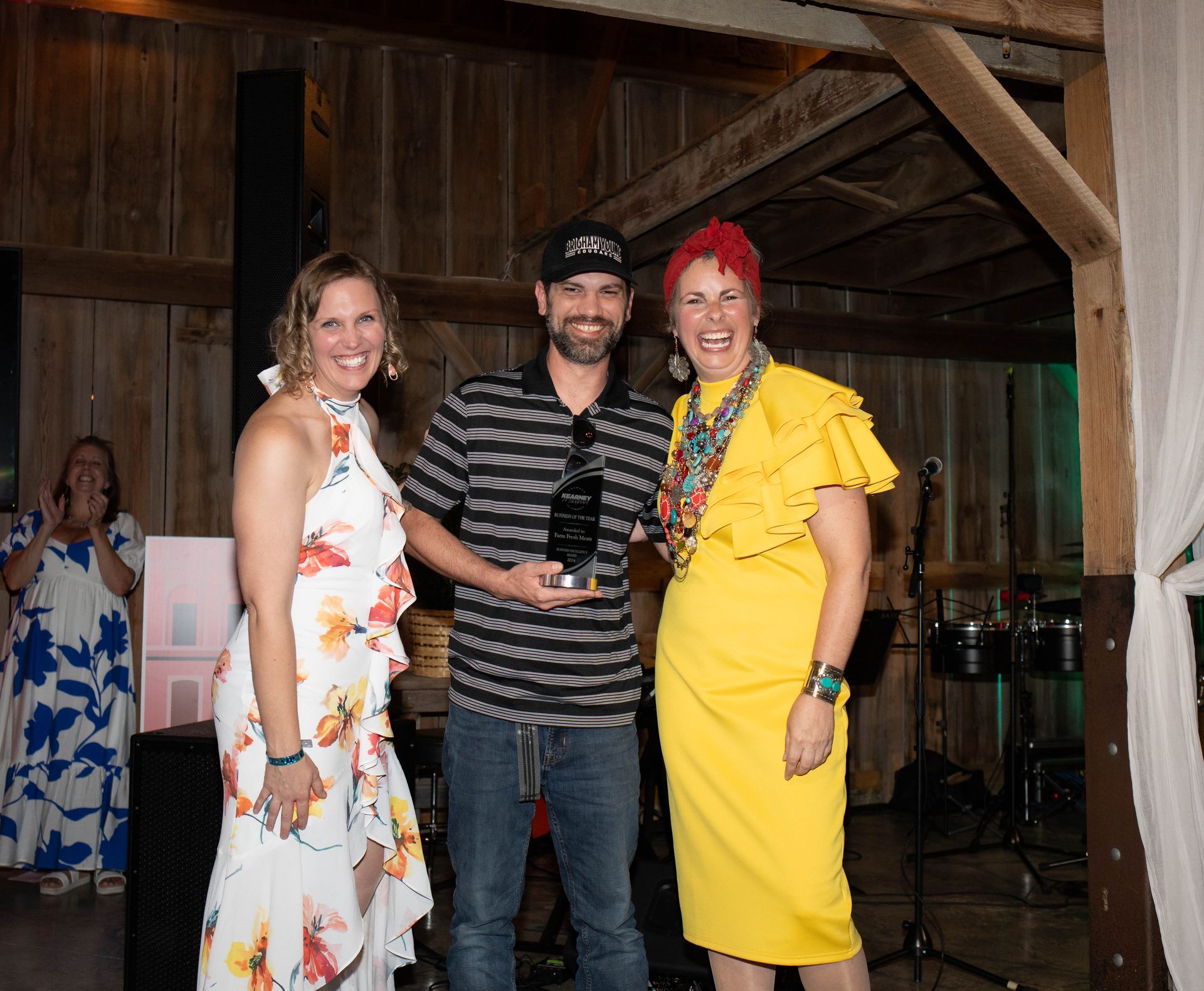 Three people smiling, posing for a photo during an award ceremony in a rustic building. A man in a hat holds an award, flanked by two women.