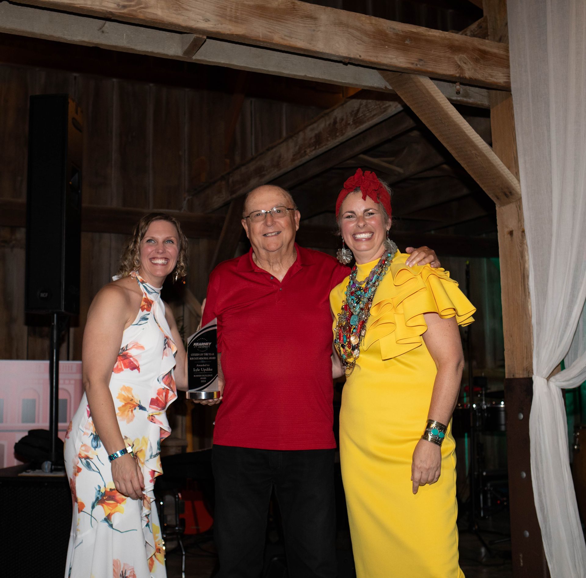 Three people smile, posing with an award in a rustic barn setting. The man in red holds the award, flanked by women in a floral dress and yellow dress.