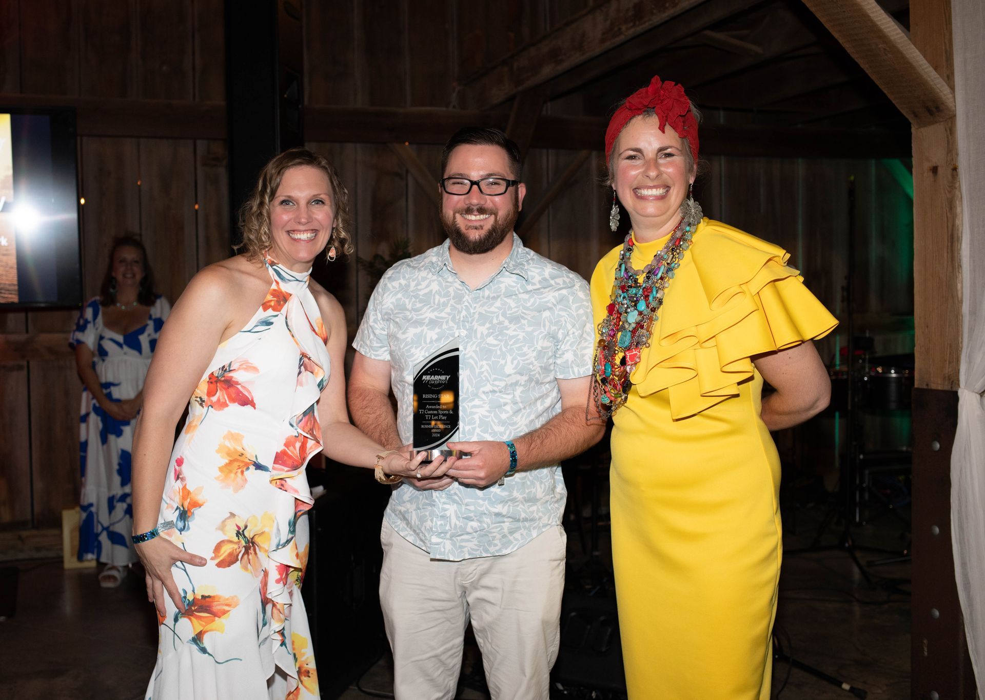 Three people at an awards ceremony in a barn. A man holds a trophy, flanked by two women in colorful dresses, smiling.
