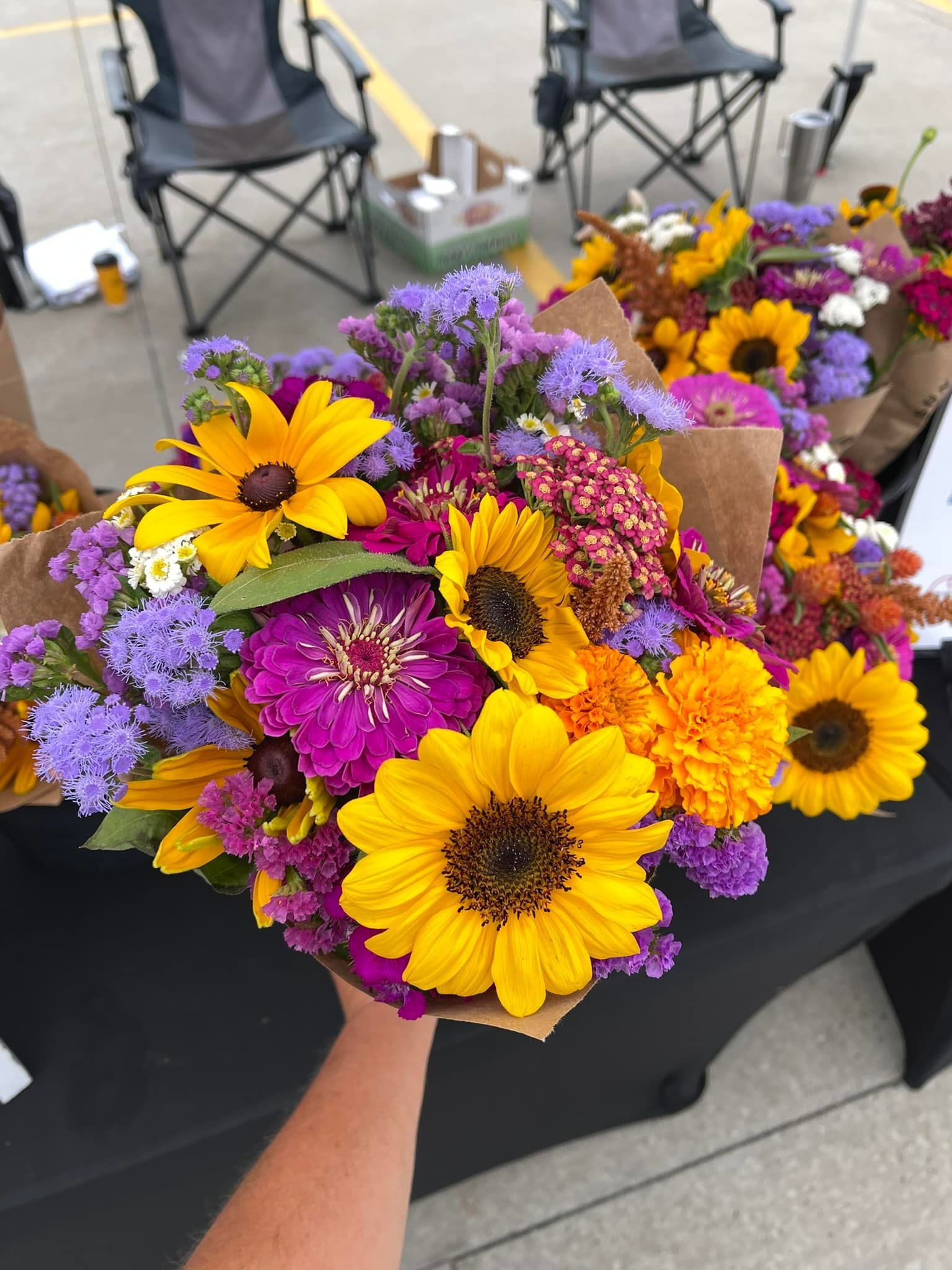Hand holding a colorful bouquet of sunflowers, zinnias, and other wildflowers at an outdoor market.