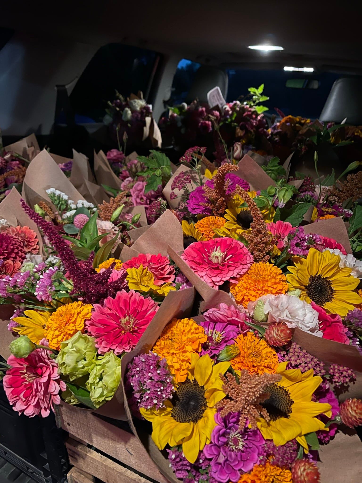 Colorful bouquets of flowers in a vehicle, likely for transport. Predominant blooms include sunflowers, zinnias, and marigolds.