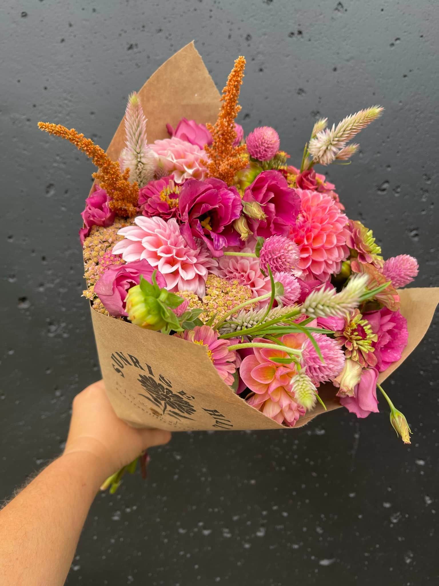 Hand holding a bouquet of pink and red flowers wrapped in brown paper against a dark background.