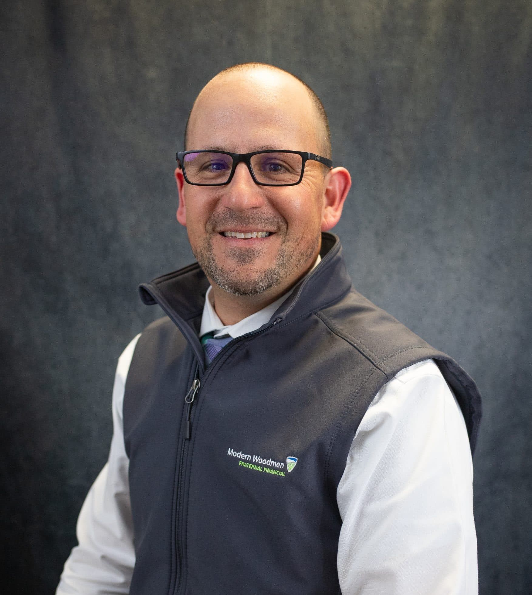 Man with glasses and a shaved head wearing a vest smiling. The vest is over a collared shirt, and he's in front of a dark gray background.
