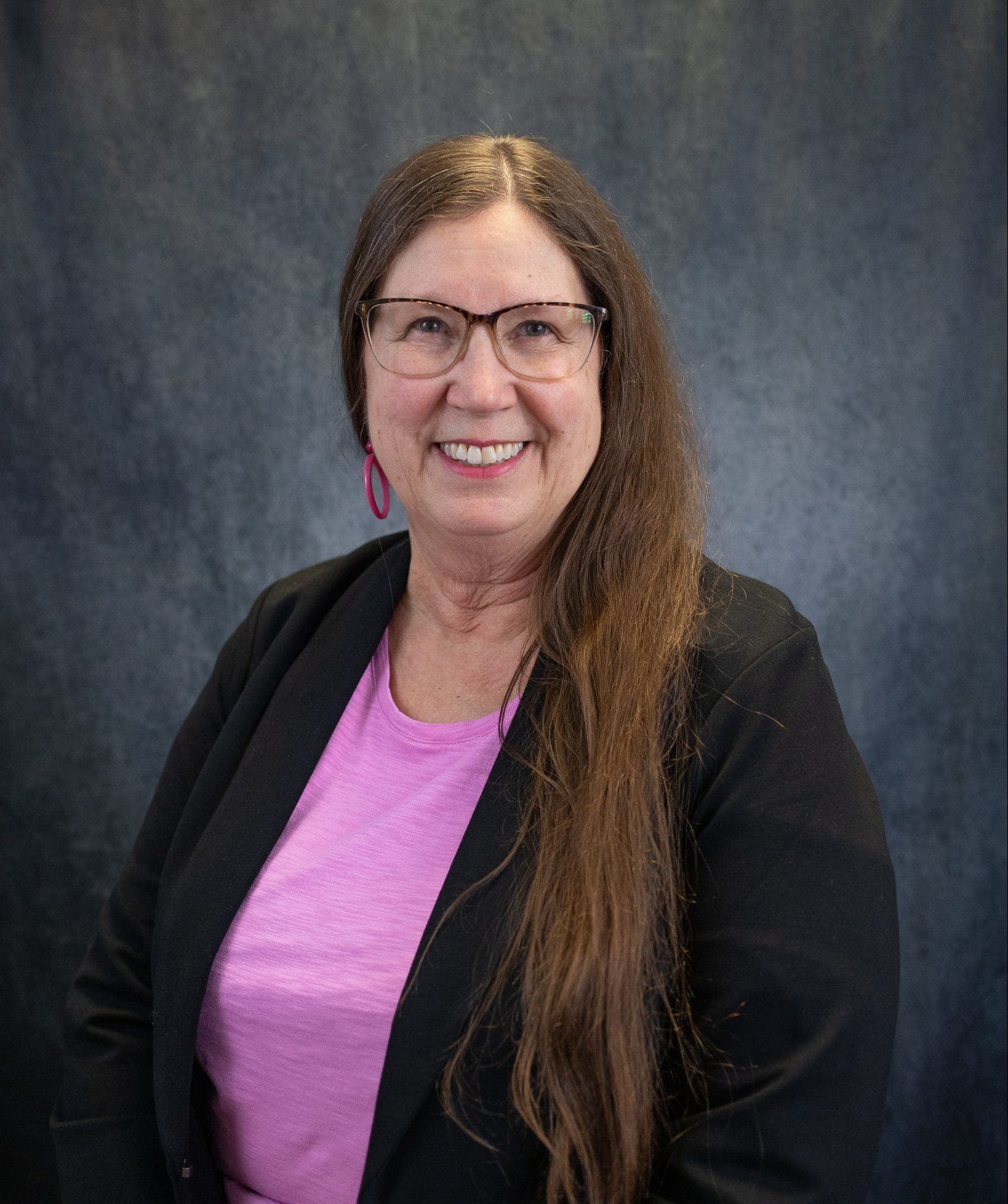 Woman with long brown hair, glasses, and pink shirt smiles at the camera, wearing a black jacket, against a dark grey background.