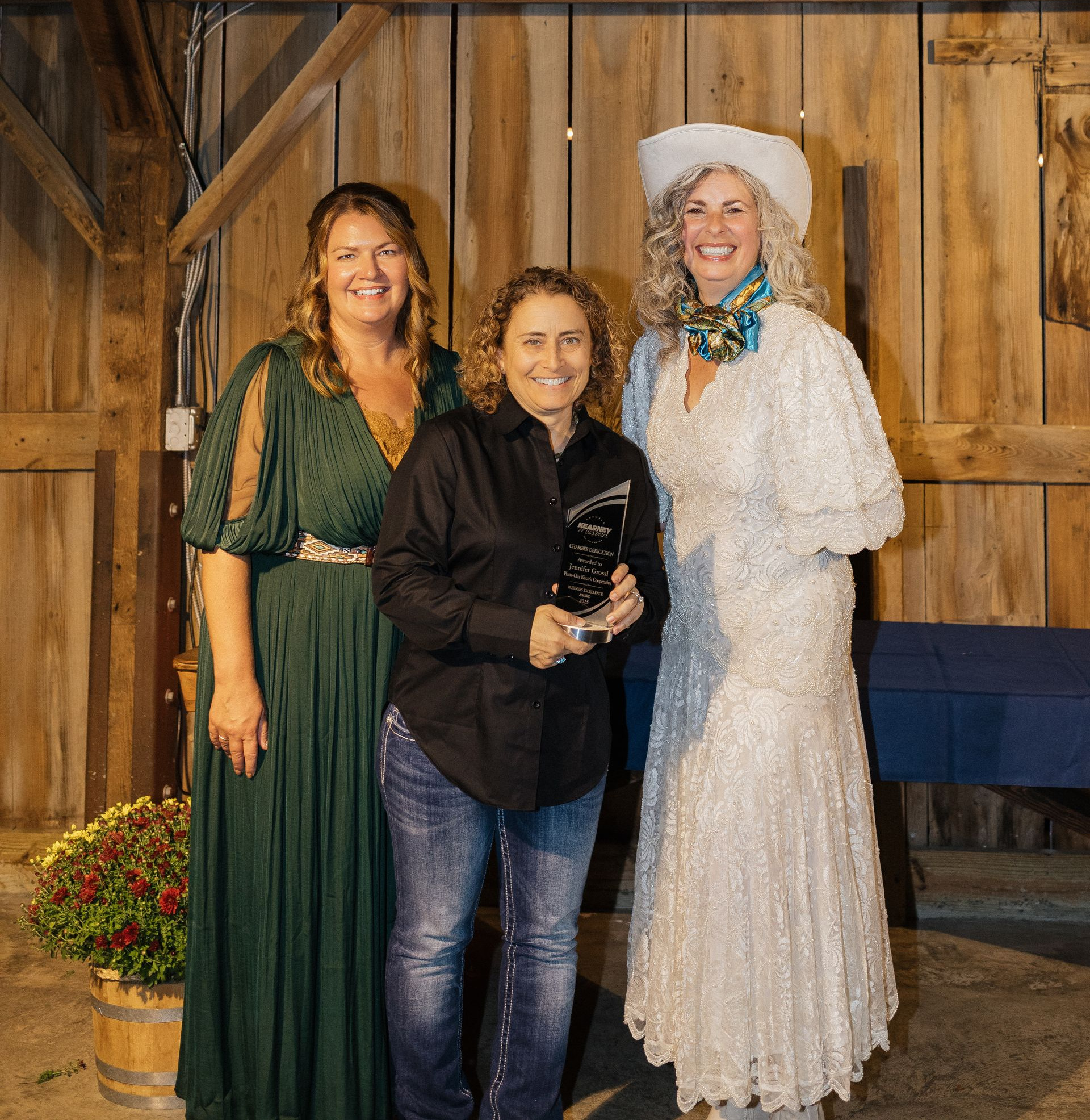 Three people smiling, posing for a photo at an event. A man in a button-down shirt holds an award. The women wear a floral dress and a yellow dress with large necklace.