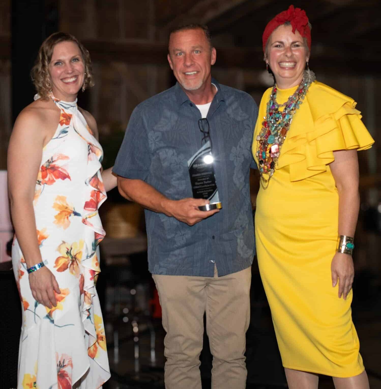 Three people smiling, posing for a photo at an event. A man in a button-down shirt holds an award. The women wear a floral dress and a yellow dress with large necklace.