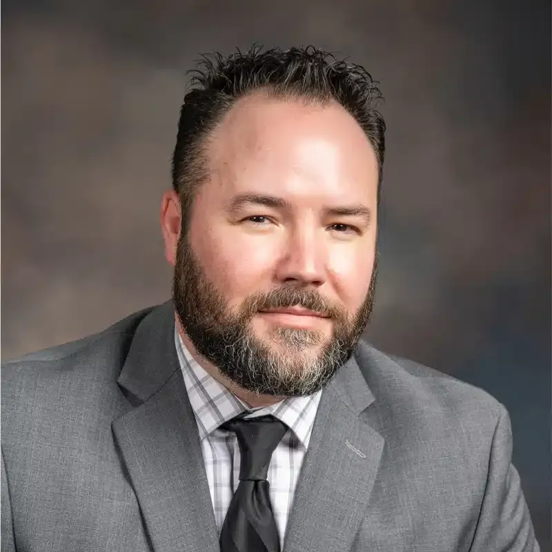 A man with a beard, wearing a grey suit and tie, smiles at the camera against a neutral background.
