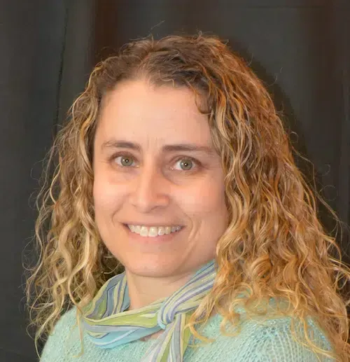Woman with curly blonde hair smiles, wearing a blue sweater and scarf, against a black backdrop.