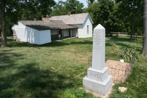 A white house and a stone tomb in a grassy area, marking the grave of someone named James. The tomb has writing on it.