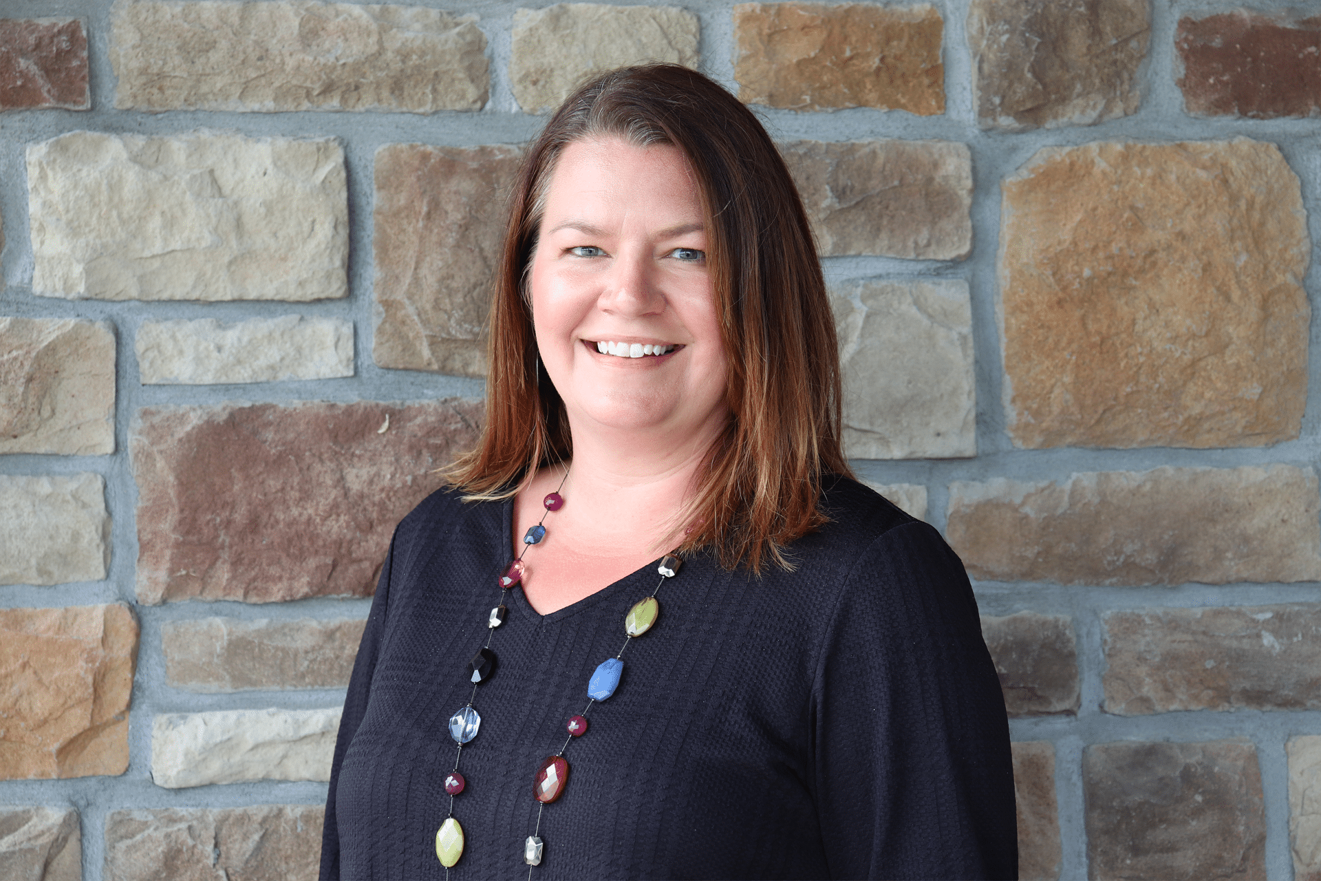 Woman with shoulder-length brown hair and a black shirt smiles in front of a stone wall, wearing a colorful necklace.