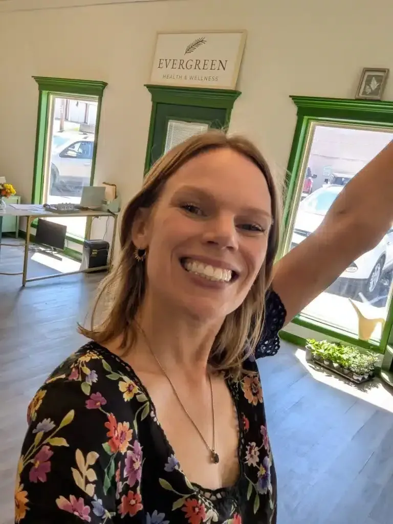 Woman with fair skin smiles at the camera, posing in front of an interior decorated with green accents. She is wearing a black floral top and has a necklace on.