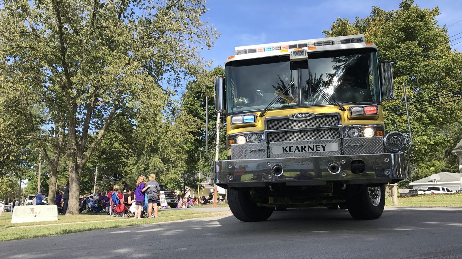 A yellow and silver fire truck drives on a road, with a crowd watching from the side, and trees in the background.