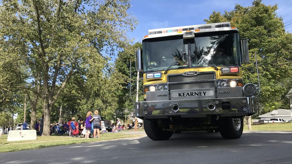 A yellow and silver fire truck drives on a road, with a crowd watching from the side, and trees in the background.