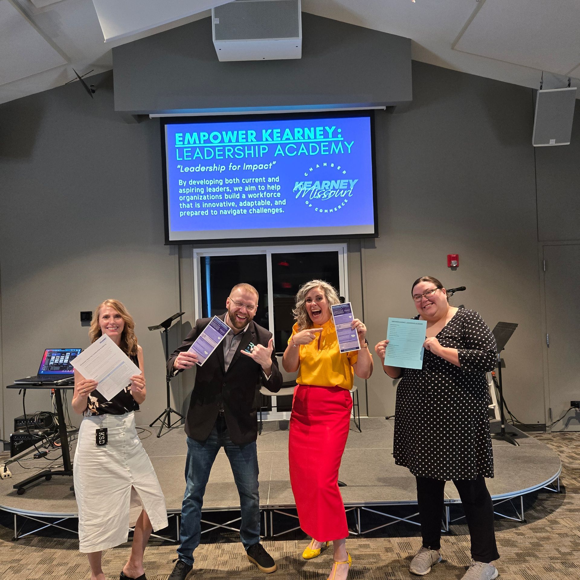 Four people smiling and holding papers on a stage in front of a screen that says 