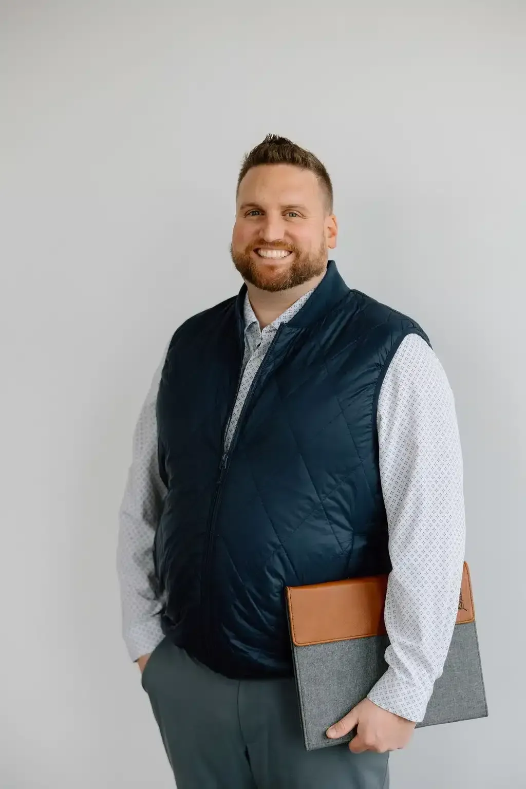 Man with a beard, wearing a vest and patterned shirt, smiling and holding a laptop case; standing in front of a white wall.