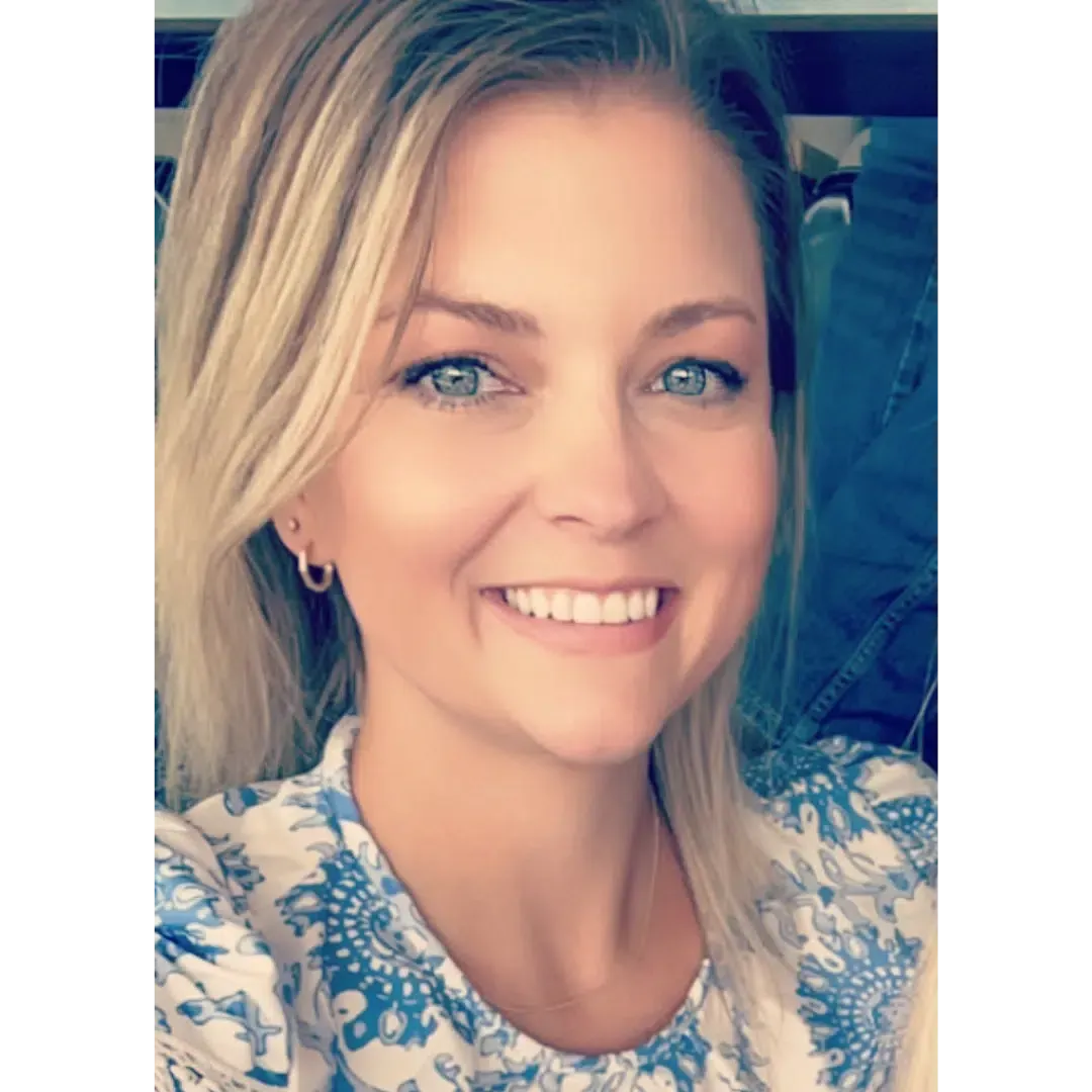 Blonde woman smiling at the camera, wearing a blue and white patterned shirt. She has blue eyes and small hoop earrings.