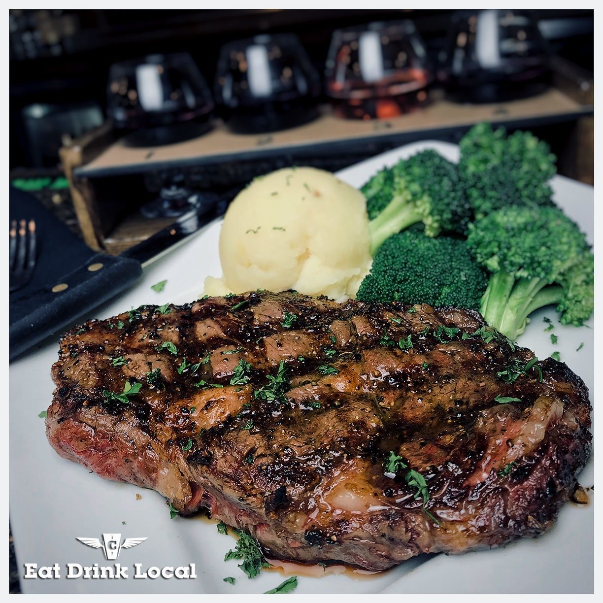 Grilled steak, mashed potatoes, and broccoli on a white plate, with wine glasses in the background.