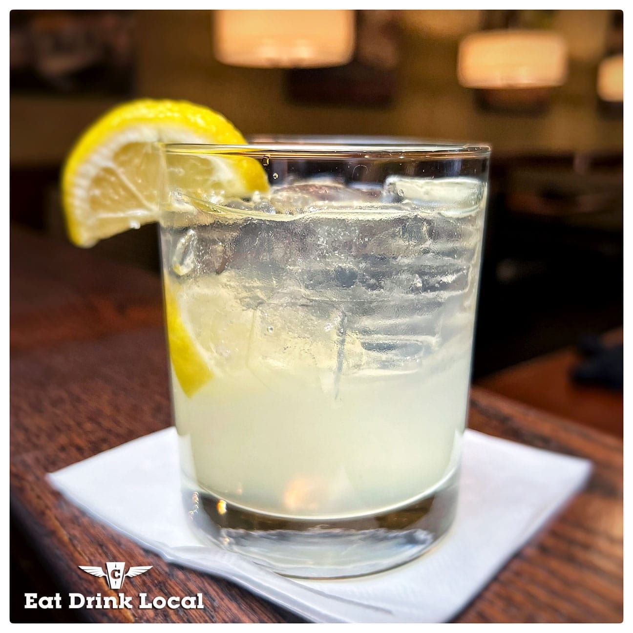 A lemon wedge-garnished cocktail in a glass filled with ice, sitting on a white napkin on a wooden bar.